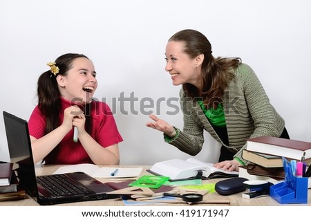 Female teacher and schoolgirl teenager at a school desk socialize animatedly, are laughing, smiling.