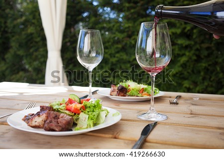 Barbecue meat served with red souse and green mixed salad. Pouring wine into glass. Royalty-Free Stock Photo #419266630