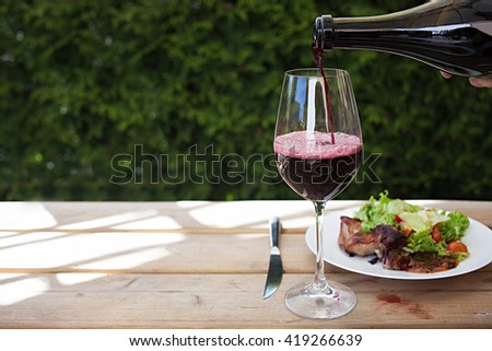 Barbecue meat served with red souse and green mixed salad. Pouring wine into glass. Royalty-Free Stock Photo #419266639