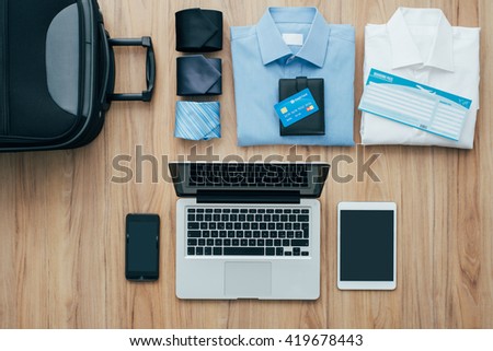 Planning a business trip: bag, formal clothing, credit cards and plane tickets on a desk with laptop, smartphone and tablet, traveling and technology concept Royalty-Free Stock Photo #419678443