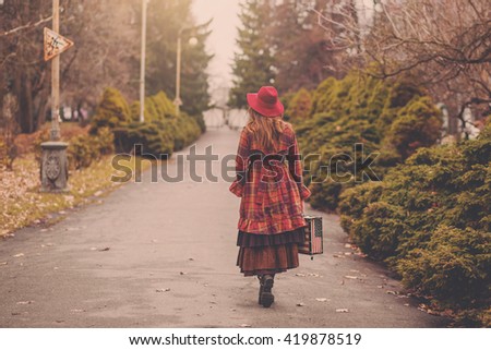 Woman in a burgundy hat and plaid winter coat is the alley e cold foggy park with suitcase in the form of American flag Stars and Stripes, English vintage