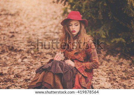 Beautiful young woman in vintage burgundy coat sitting on carpet of autumn leaves in the cold park with suitcase in the form of American flag Stars and Stripes, English vintage