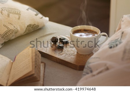 Hot coffee in a white mug with a nice smoke on the wooden coaster, dark chocolate pieces and an open book in bed. Grey sheet and soft pillow background. Morning light. Toned picture. Vintage look.

