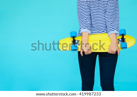 girl holding a skateboard, wearing casual shirt and jeans, view from back isolated on blue background. Hipster lifestyle concept