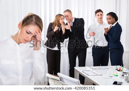 Businesspeople Gossiping Behind Stressed Female Colleague In Office Royalty-Free Stock Photo #420085351