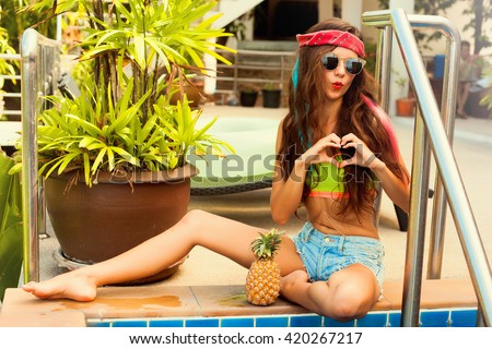 Summer portrait of pretty brunette girl,kissing lips,having fun with pineapple.Singing with closed eyes and smiling.Casual style,bright makeup,red lips.street background, not isolated.red bandana