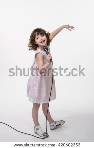 Laughing singer with black microphone in her hand, the other one is rised. Lovely curled brunette hair and cute pastel pink dress are very cool for girlish style. Positive emotions of a child.