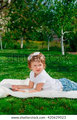 baby girl dressed in white polo and jeans, barefoot lying with tablet on the fur blanket in the park with blossoming trees in the background. Girl Using a Tablet. 