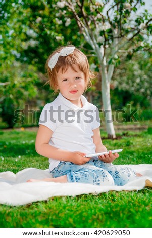 Little baby girl dressed in white polo and jeans, barefoot sitting with tablet on white fur blanket in park with blossoming trees in the background. Girl Using a Tablet. looking at the camera