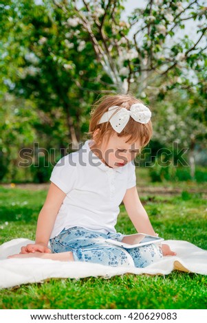 Little baby girl dressed in white polo and jeans, barefoot sitting with tablet on the white fur blanket in park with blossoming trees in the background. Young girl getting into learning on tablet