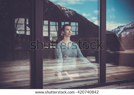 Young beautiful woman meditating while practicing yoga with mountain view in the window reflection. Freedom concept. Calmness and relax, woman happiness. Toned picture
