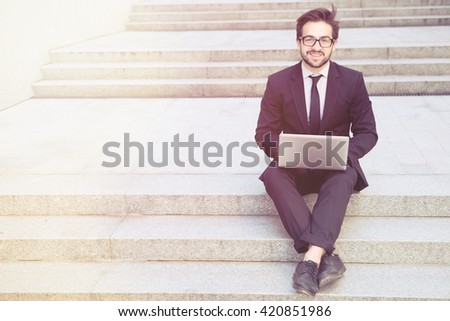 Toned image of confident and executive businessman looking at camera while using laptop computer in city centre. Smiling freelance man working on street.