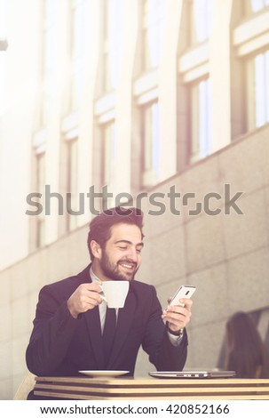 Toned picture of happy businessman drinking coffee or tea while working in restaurant or cafe. Freelance man using mobile or smart phone.