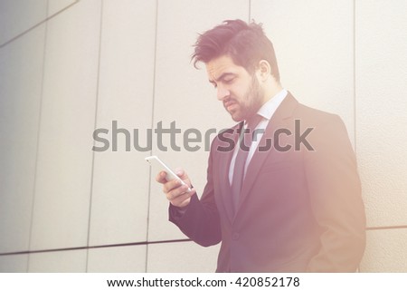 Toned portrait of young handsome businessman in black business suit looking at mobile or smart phone while standing near office building in city centre.