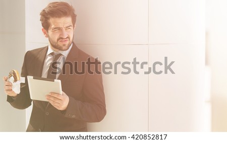Toned picture of young businessman in business suit looking away and posing with tablet PC while eating junk food on street.