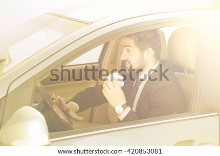 Toned picture of young businessman eating junk food while driving to work to office. Handsome man in black business suit looking to traffic jam. 