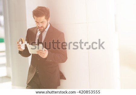 Toned picture of businessman standing near office building and looking at tablet PC. Handsome man eating junk food on street.