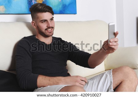 Attractive young man taking selfie with smartphone on white sofa. Indoor.