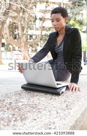 Beautiful black business woman sitting on a stone bench in the financial city, using a laptop computer, thoughtful outdoors. Professional african american woman using technology, working lifestyle.