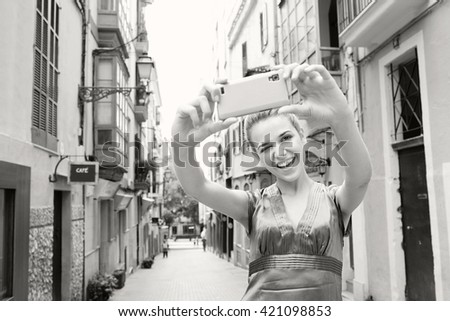 Black and white portrait of beautiful tourist woman holding up a smart phone, taking selfies photos, in a destination city street, holiday exterior. Woman using technology, travel lifestyle.