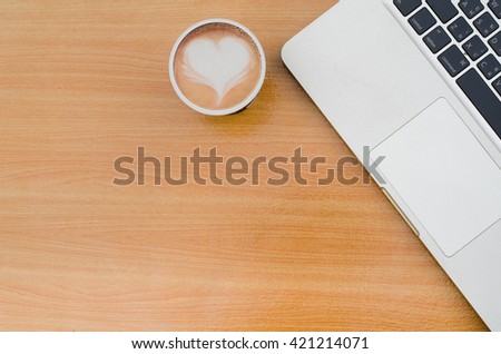 Selective focus of coffee latte art heart with blurred notebook (laptop) on workspace of wooden table. Concept for office workplace, freelance, coffee time, blogger, social, programmer, background