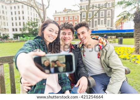 Three friends taking a selfie together. They are sitting on a bench at park, two boys and a girl, wearing autumn clothes. They are happy, smiling and enjoying time together.