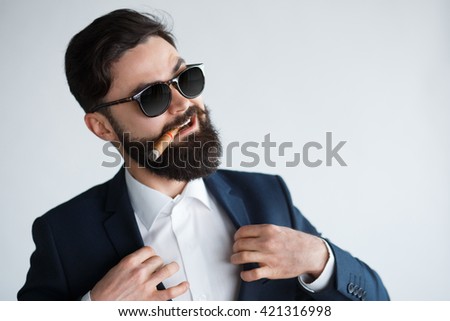 young confident businessman smoking a cigar and fixing his black suit with confidence on white background. Arrogant rich bearded man in sunglasses Royalty-Free Stock Photo #421316998