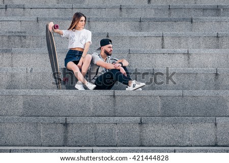 Young stylish couple dressed in hipster style on concrete stairs into the summer city. Stylish couple in the urban landscape. Sunny portrait of young stylish couple in the urban landscape.