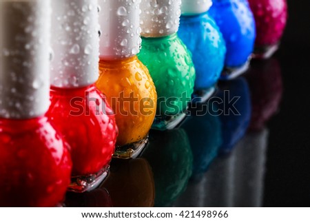 Group of bright nail polishes sorted like a rainbow. Bottles with water drops are standing in a row on black stone background with reflection. Beauty concept.