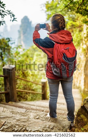 Young female tourist with red backpack standing on stone stairs among green foliage and taking photo of beautiful mountain view, the Zhangjiajie National Forest Park, Hunan Province, China.