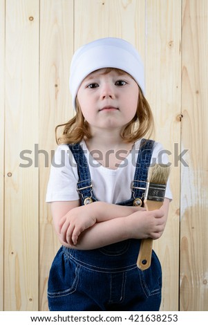 Little girl in the form of a painter with a brush in his hands posing on the background of wooden wall