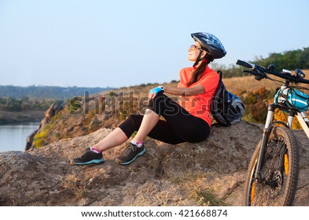 Adult attractive female cyclist sitting on the rock with closed eyes and enjoying the evening sunlight.
