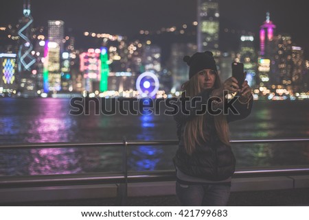Young attractive blonde woman makes self portrait using mobile phone with view over Victoria Harbor at night in Hong Kong, China. Teenage girl makes picture.