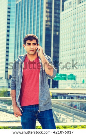 East Indian American college student traveling in New York, wearing red v neck shirt, gray hooded sweatshirt, standing in front of business district, talking on cell phone. Instagram filtered effect.
