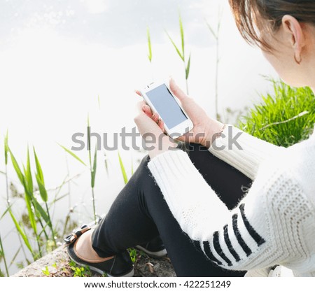 Beautiful young woman using mobile phone, sitting on the river bank. Summer photo. Coastal view.