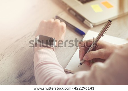 Close up of woman hands  with smart-watch writing business ideas in notes near laptop.
Sun glare.