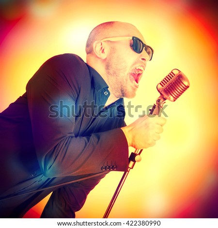 A man in a gray jacket, bow tie and sunglasses with a microphone in hand, singing on the bright abstract background. Toned