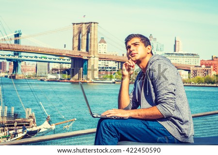 East Indian American student traveling, studying in New York, sitting by river, working on laptop computer, tilting head, hand touching cheek, looking up, thinking. Brooklyn bridge on background.