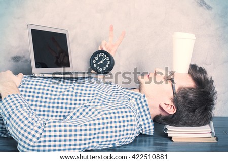 Sleepy caucasian businessman with blank laptop and coffee cup on forehead, lying on office desk and showing peace sign. Mock up