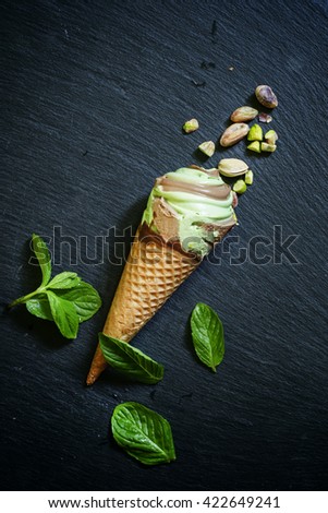 Mint pistachio ice cream with chocolate in waffle cone, scattered pistachio and mint leaves, dark stone background, selective focus