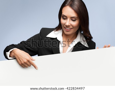Portrait of happy smiling young businesswoman in black suit, showing blank signboard with blank copyspace area for slogan or text, over grey background