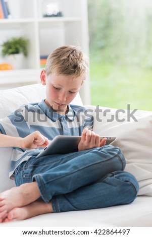 At home, 8 years old boy having fun by playing on a tablet, he is sitting on a white couch in the living room