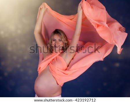Beautiful blond  pregnant woman dressed in pink with waving tissue looking at camera, isolated on dark blue background