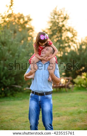  A happy family. Daughter kisses her mother whereas smiling father is carrying his daughter on his shoulders at nice sunny background.