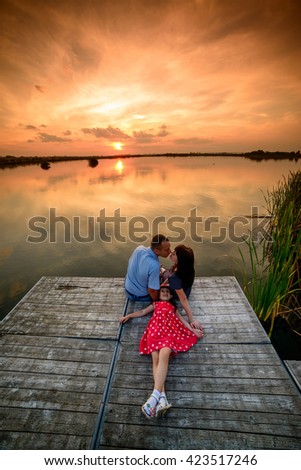 A friendly family is lying on the wooden bridge at the sunset whereas the golden sun is reflecting in water adding cozy atmosphere of the picture