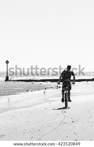 Person riding a bicycle on the beach on hard packed sand next to the water. Viewed from behind. Black and white. High key image. Copy space.