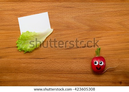 Mr. Radish is smiling and looking at the blank card, next are the lettuce on wooden table. Close-up view from above