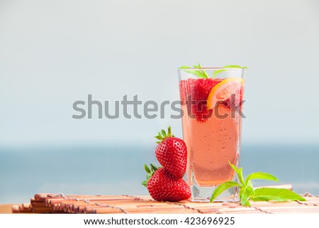 Glass of strawberry lemonade with pieces of strawberry, lemon and fresh mint. Closeup of mojito with beach on background. Fresh summer bright cocktail.