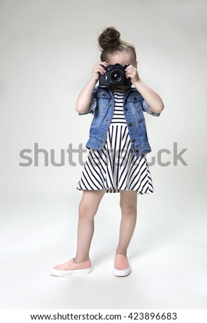  ?eautiful smiling little girl with a camera taking pictures in a striped dress and denim jacket on a light background in studio