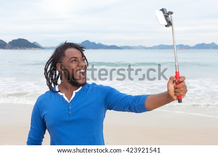 African american guy with dreadlocks at beach taking picture with selfie stick with ocean and sky in the background